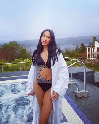 Woman in a black bikini and white cover-up standing by a pool with a scenic background